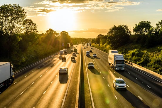 Cars and lorries driving on a motorway with trees and the sun setting in the background.