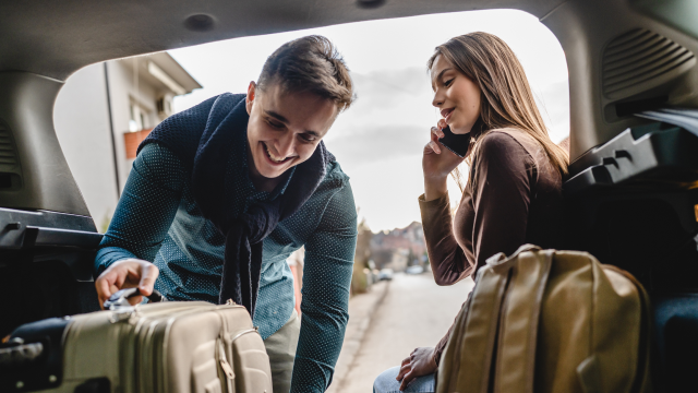 A man smiles, taking a suitcase out of a car, while a woman looks on and talks on the phone.