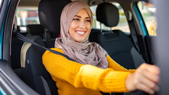 A woman in a yellow jumper sits in the driving seat of a car, smiling and with her hands on the steering wheel.