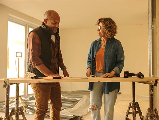 Home improvement loans A man and a woman working on a home improvement project. They're in a room with protective coverings on the floor, standing behind a plank of wood resting on two sawhorses. Tools are placed on the plank of wood.